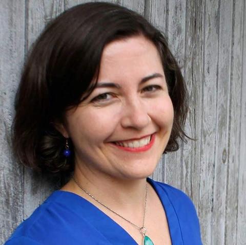 Color headshot of RJ Taylor, a fair-skinned woman with dark, lightly wavy hair parted on the side and worn in a chin-length bob without bangs. She wears a silky blue V-neck blouse, dangly earrings with what looks like a single lapis lazuli bead each, a necklace with a large teardrop pendant, and coral lipstick. She is turned slightly away from the camera, her back against a wall of weathered wood. She smiles warmly at the camera, a smile that goes all the way to her brown eyes.