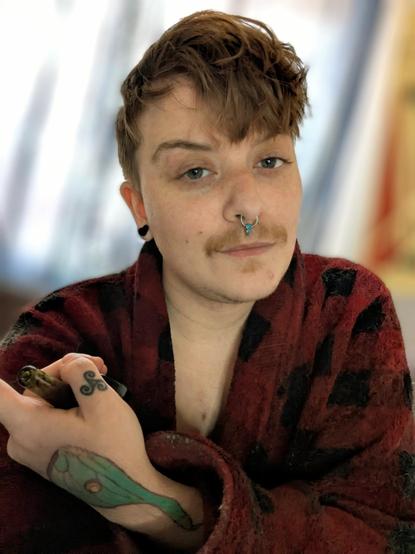 Trans masc fox in a red and black checkered fleece robe holding a glass pipe with a tattooed hand. He has freckles, short red hair and a mustache