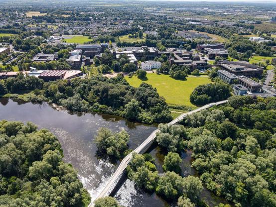 Aerial photograph of the University of Limerick campus, showing buildings nestled amongst mature trees and a large river spanned by a curved pedestrian bridge