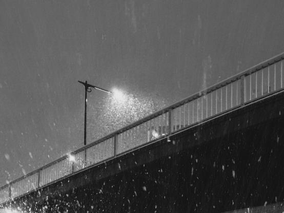The image depicts a black and white scene of a bridge at night with street lamps illuminating the area. Snow or rain is falling, visible in the light, creating an atmospheric effect. The lighting and shadows add a dramatic contrast to the scene.