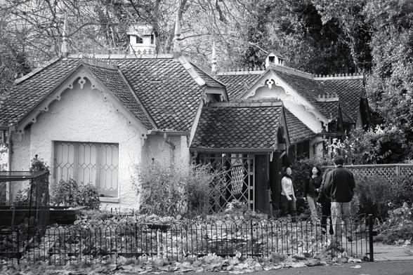 Black and white. Four people, one man and three women, are standing or walking in the front garden of Duck Island Cottage in St James's Park. It is a Swiss chalet-style cottage with two small buildings connected by a covered walkway that crosses a bridge, with tiled roofs, decorated gables, several small spires, and a tall white chimney stack topped with what looks like a small bird house. Seen in colour, it looks somewhat like a gingerbread cottage. It is often topped off by a grey heron enjoying the view over the lake behind the cottage.