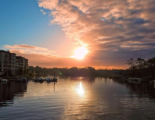 Bright, golden sunrise casts glowing light across a waterway below and colorful reflections upon cloud formations in the blue morning sky.