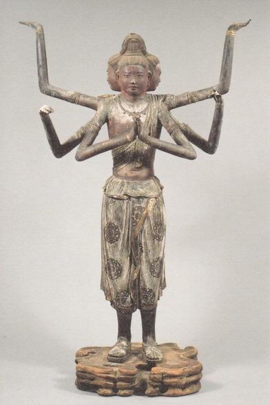 a tall  realistic sculpture of a slender figure with a 4-sided head and six arms, the arms held out in different angles, wearing baggy short pants with patterns on them, facing the viewer, against a grey background