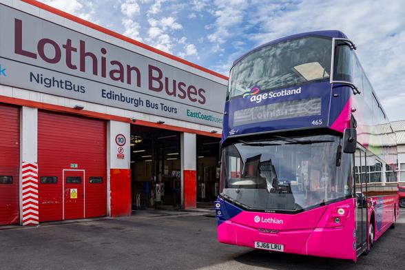 The image shows a bright, modern double-decker bus parked outside the Lothian Buses depot. The bus features a vibrant purple and pink colour scheme with the “Age Scotland” logo visible on the front above the windscreen. It is branded “Lothian” with fleet number 465 and bears the registration plate SJ66 LRU. The depot building, mainly red and white, has large roller shutters, one of which is open. Above the doors, a large sign reads “Lothian Buses”, with additional services listed, including NightBus, Edinburgh Bus Tours, and EastCoastbuses. A small road sign indicates a speed limit of 10 mph.

Photo by Lothian Buses ltd.