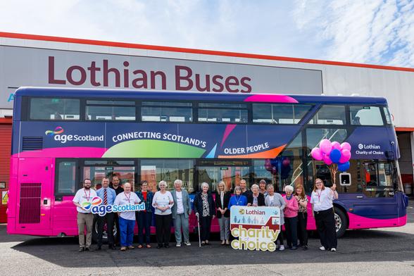 The image also features a vibrant double-decker bus parked in front of a large building with “Lothian Buses” signage. The bus sports a bold design, with a bright magenta lower half and a navy blue upper section. The sides display the message “Age Scotland – Connecting Scotland’s Older People”, with multicoloured abstract shapes. In front of the bus stands a diverse group of smiling people holding signs for “Age Scotland” and “Lothian Charity of Choice”. Some are holding pink and purple balloons. The scene takes place outdoors under a partly cloudy sky, suggesting a daytime event.

Photo by Lothian Buses ltd.
