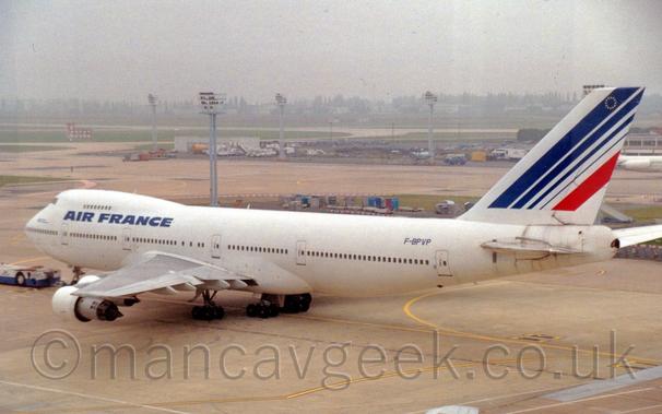 Side view of a very large, white, 4 engined jet airliner being towed from right to left.
The plane is almost entirely white, with blue "Air France" titles between the upper and lower decks at the front of the fuselage, and a series of blue diagonal stripes (and a single red stripe) on the tail.
The blue registration "F-BPVP" is on the upper rear fuselage, just in front of the tail.
A white and blue tug is attached to the nose, pulling the plane across the frame.
Tall lighting poles are scattered around the background, with grassed aeras and vehicle parks separating taxiways, slowly vanishing into haze in the distance.