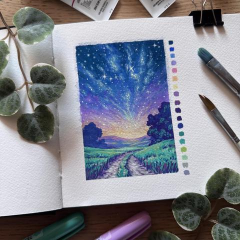 A photo of an open sketchbook placed on a wooden desk and surrounded by paint tubes, paint brushes, acrylic markers, and leaves for decoration. The sketchbook  features an open rural landscape under a twilit sky with a galaxy. A path leads from the foreground towards a few trees and softly rolling hills in the background. A colour swatch is painted next to the scenery.