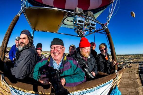 Several photographers in a balloon looking into the camera. One is smiling broadly. The balloon is well off the ground. In the background another hot air balloon higher up in the blue sky.