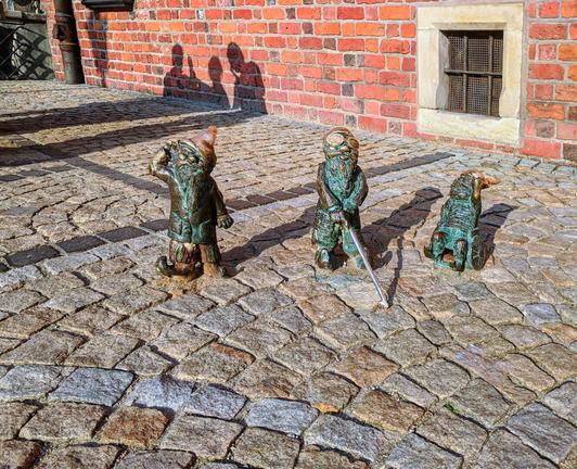Three bronze statues of Wrocław Dwarfs stand on a cobblestone street. The dwarf on the left, representing deafness, wears a pointed hat and puts a hand to his ear, looking upwards. The dwarf in the middle, representing blindness, holds a white cane. The dwarf on the right is in a wheelchair. A red brick building with a window is visible in the background. The shadows of people are cast on the building.