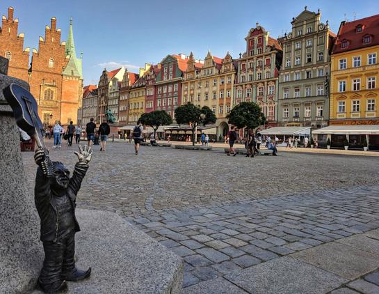 A bronze statue of a Wrocław Dwarf stands on a cobblestone street. The dwarf is holding a guitar above its head, its arms outstretched. In the background, there's a bustling market square with colourful buildings, people walking around, and a church tower visible on the left.