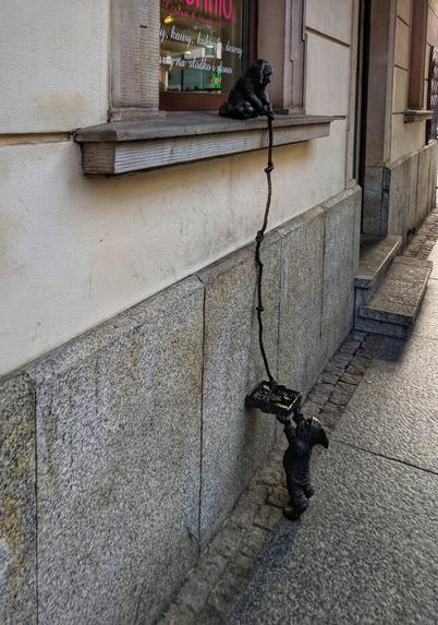 Two bronze statues of Wrocław Dwarfs are positioned on the exterior of a building. One dwarf sits on a window ledge, while the other is standing on the street, seemingly pulling the first dwarf up with a rope. The building's exterior is made of stone and features a window with text partially visible above it.