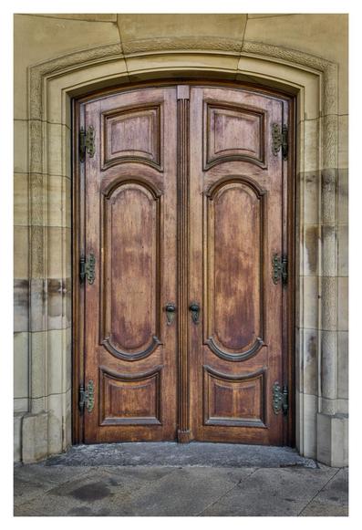 The picture shows a large, double-leaf wooden door with a rustic, brown finish. The door has two elongated, oval recesses in the centre, which are surrounded by simple, rectangular frames. There are solid, wrought iron hinges on the sides of the door, which have a slightly patinated appearance. A round, black door knob is attached to both door leaves. The door is set in a stone arch frame made of light beige, textured stone. The floor in front of the door is covered with grey cobblestones. The entire scene conveys an impression of age and solidity.