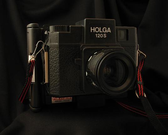 Holga 120S camera with Polaroid back against a black background. Black gaffers tape is seen in various spots holding bits together. A red and black camera strap is clipped to either side.