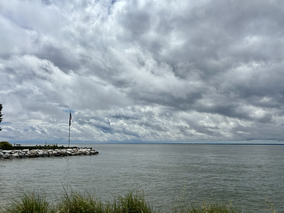 dramatic, overcast sky. The foreground features some tall grass along the water’s edge, adding a natural texture. A stone jetty extends into the water on the left side, topped with a flagpole flying the American flag. The water is calm, reflecting the gray tones of the sky above.