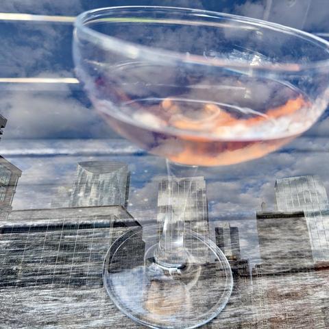 A cocktail glass filled with a pink drink is displayed in the foreground, reflecting a cloudy sky and city skyscrapers in the background. The image captures an urban setting with a mix of glass and concrete structures.