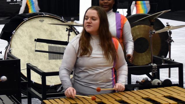 A performer playing a mallet percussion instrument during a music performance, wearing a silver long-sleeve outfit with colorful accents. A large bass drum and other percussion instruments are in the background.