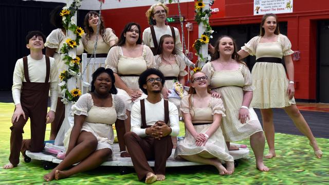 A group of eleven performers poses together, wearing matching cream-colored dresses and brown accents. They are arranged in a playful manner, with some seated and others standing, smiling joyfully. The background features a colorful floor resembling grass.
