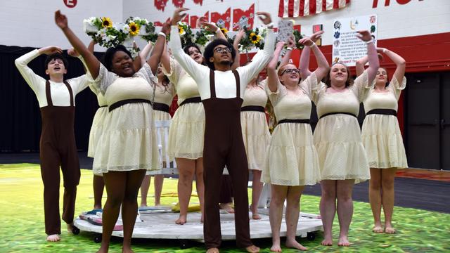 A group of performers is on stage, dressed in short, cream-colored dresses and brown suspenders, celebrating with raised arms. The scene includes floral decorations, and they appear to be part of a theatrical or dance performance in a school gymnasium.