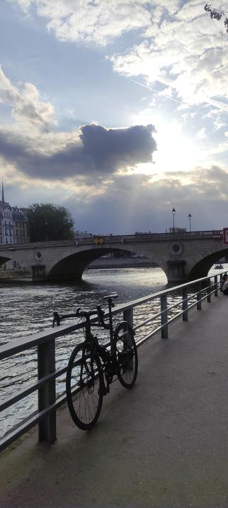 Un vélo noir appuyé contre une rambarde métallique, sur un quai parisien. Le ciel est chargé, traversé par des éclats de lumière. En arrière-plan, un pont, la Seine, et la ville. L’ambiance est tendue et belle, comme un souffle repris en plein cœur du mouvement. Bikechaser en pause, mais le regard toujours allumé.
