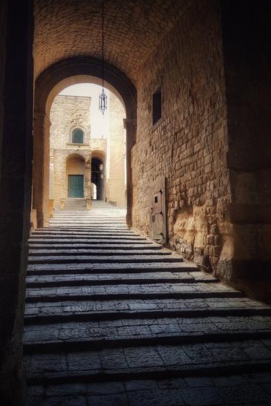 A sunlit, arched stone passageway with a long, wide set of stone steps leading upwards. The walls are made of rough-hewn stone, and a green door is visible at the top of the steps. Light streams in from the opening at the end of the archway, illuminating the steps and the building beyond.