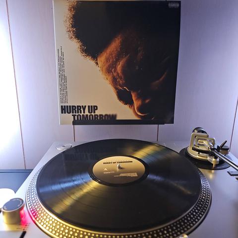 A vinyl record sits on a turntable. Behind the turntable, a vinyl album outer sleeve is displayed. The front cover shows The Weeknd's face partially cut off. He has his eyes closed and his forehead is covered in sweat .
