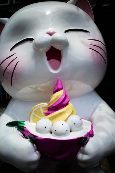 The image shows a large, shiny white cat figure with a wide open mouth, as if joyfully laughing. The cat holds a colorful ice cream dish, featuring swirled pink and yellow ice cream, round white balls resembling mochi, and a slice of lemon on the rim.