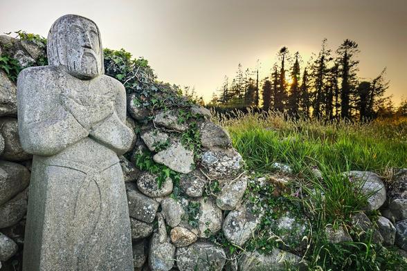 A stone figure with folded arms stands on the left, next to a stone wall covered in ivy and moss. To the right, a grassy field leads to a line of tall trees, with the sun setting behind them, casting a warm glow.