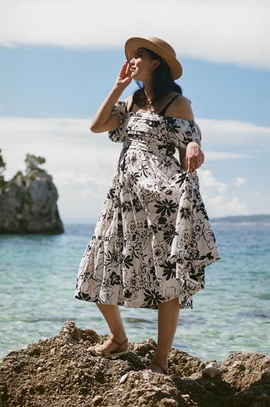 A beautiful woman with dark hair, wearing a gold necklace, a straw hat, and a long white and black summer dress, standing on a rock in front of the Mediterranian sea.