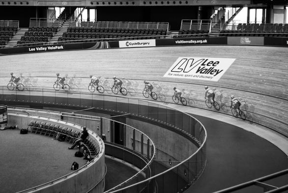 A black and white photograph shows an indoor cycle track, the Lee Valley VeloPark, with a group of cyclists racing. The track curves steeply, and the cyclists are captured with a motion blur effect, creating a sense of speed. Banners with "Cycle Surgery" and "visitleevalley.org.uk" are visible along the track's edge. In the background, there are rows of empty seats. The overall scene captures the dynamic energy of a cycling event in a modern velodrome.