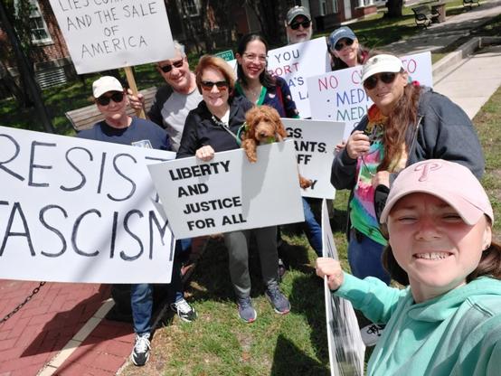 A group of 8 protesters and a young dog are standing together for a selfie as they hold protest signs.  Examples of the signs are, "Resist Fascism," and "Liberty and Justice for All."