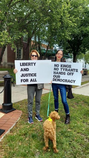 Two women are standing side-by-side with protest signs.  The signs read, "Liberty and Justice for All," and "No Kings, No Tyrants, Hands Off Our Democracy." A furry, tan dog is on the grass in front of them.