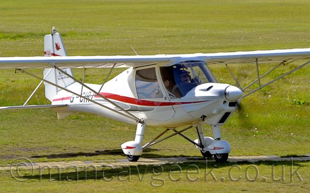 Side view of a white, high-winged, single-engined light aircraft taxiing from left to right on grass.
The plane has a red and grey stripe running along the body, and the black registration "G-CIRZ" on the tapered rear fuselage.
2 men can been seen in the cockpit through the large windows.
Green grass fills the rest of the frame.