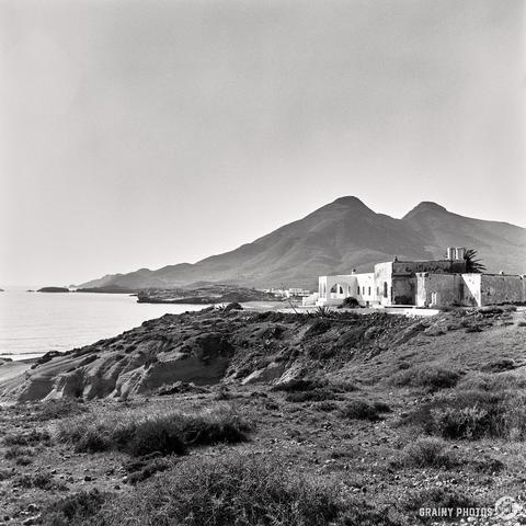 Captured using a Mamiya C220, this black and white image showcases a rocky Cabo de Gata coastline with several white-washed buildings near the shore. The scene is set against mountains under a clear sky.