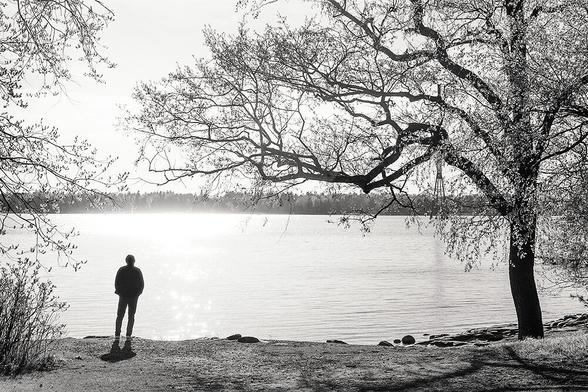 A silhouetted figure stands by a tranquil sea, framed by trees, reflecting soft sunlight in a serene black-and-white scene.