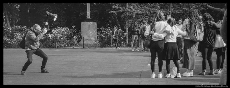 A photographer crouches with a large camera setup in preparation for taking a photo of a group of people on the right, arranged in a semicircle.