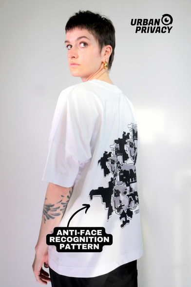 Das Bild zeigt die Rückseite einer Person, die ein schwarzes T-Shirt mit einem auffälligen Design trägt. Das T-Shirt hat einen großen, weißen und grauen "ANTI-FACE RECOGNITION PATTERN" auf der Rückseite, der aus pixelartigen Formen und Zahlen besteht, die an eine digitale Grafik erinnern. Der Text "URBAN PRIVACY" ist sowohl auf dem T-Shirt als auch oben rechts im Bild zu sehen. Die Person hat kurzes, dunkles Haar und trägt goldene Ohrringe. Der Hintergrund ist ein einfacher, neutrales Grauton, der den Fokus auf das T-Shirt lenkt.