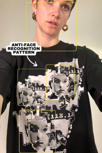 Das Bild zeigt eine Person von hinten, die ein weißes T-Shirt mit einem großen, abstrakten, schwarzen und grauen Muster auf der Rückseite trägt. Das Muster wird als "ANTI-FACE RECOGNITION PATTERN" bezeichnet, was darauf hinweist, dass es dazu gedacht ist, Gesichtserkennungssysteme zu verwirren. Die Person hat kurzes, dunkles Haar und trägt goldene Ohrringe. Auf ihrem linken Arm ist ein Tattoo sichtbar. Im oberen rechten Bereich des Bildes befindet sich das Logo "URBAN PRIVACY". Die Person steht vor einem neutralen, hellen Hintergrund.