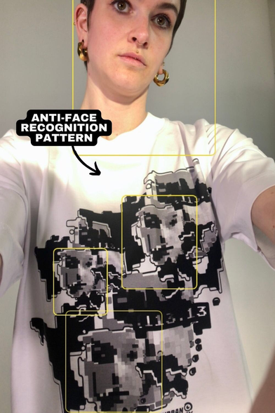 Das Bild zeigt eine Person von hinten, die ein weißes T-Shirt mit einem großen, abstrakten, schwarzen und grauen Muster auf der Rückseite trägt. Das Muster wird als "ANTI-FACE RECOGNITION PATTERN" bezeichnet, was darauf hinweist, dass es dazu gedacht ist, Gesichtserkennungssysteme zu verwirren. Die Person hat kurzes, dunkles Haar und trägt goldene Ohrringe. Auf ihrem linken Arm ist ein Tattoo sichtbar. Im oberen rechten Bereich des Bildes befindet sich das Logo "URBAN PRIVACY". Die Person steht vor einem neutralen, hellen Hintergrund.