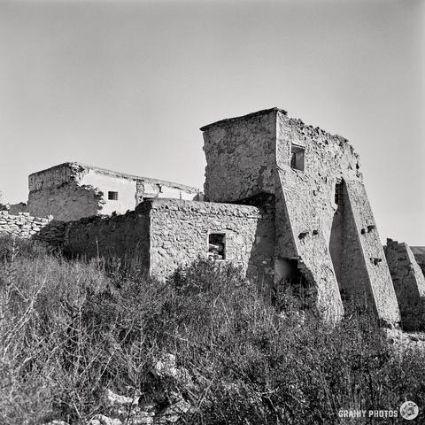 A black and white photo, captured with a Mamiya C220 on Kentmere 100 film, showcases an abandoned stone building with crumbling walls and reinforced beams. It is surrounded by wild grass and vegetation under a clear sky.
