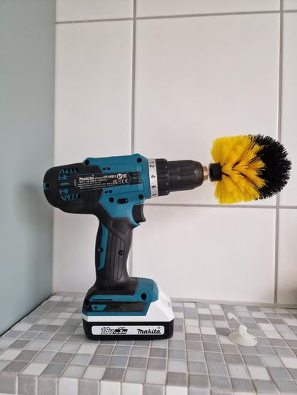 Electric handheld drill with cleaning brush mounted, photographed on white and grey mozaiek times and great white tiles In the background