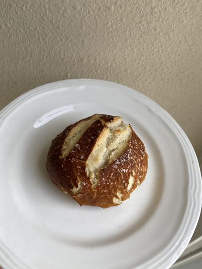 A closeup of a freshly baked pretzel roll. The skin is dark brown and there are two large gashes on the top revealing the white bread interior. All over the surface are large salt grains.