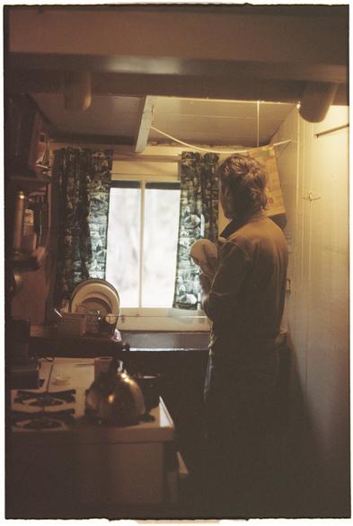 A film photo of a person with their back turned to the camera standing in a small kitchen inside a fish camp. There is a stove with a tea kettle and a window with dark green curtains. The photo is overall dark and moody with brown and yellow/orange hues and faded shadows.
