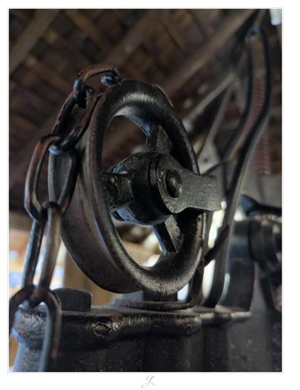 Close-up of a metal pulley with a chain running over it, shot from an angle below. The pulley has three spokes, it is attached to the heavy metal frame of a large press. All parts are lacquered black. The picture has a quite dark mood, just in the background there are some blurred light sources.

AI disclaimer: Using my work, its meta data, written or derived description to create media with or train AI based systems is prohibited.