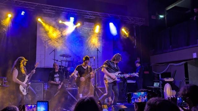 The image depicts a live music performance on a stage with a band playing. The stage is illuminated with vibrant blue and yellow lights, creating a dynamic atmosphere. The band consists of five members, each playing different instruments. From left to right, the first member is playing an electric guitar, wearing a black sleeveless shirt and a cap. The second member is seated at the drums, wearing a black t-shirt with a graphic design. The third member is playing a bass guitar, wearing a black t-shirt. The fourth member is playing an electric guitar, wearing a black leather jacket. The fifth member is playing a keyboard, wearing a dark shirt. The background features a large screen and stage equipment, with smoke effects enhancing the visual impact. The audience is visible in the foreground, with some members capturing the performance on their phones. The overall scene conveys an energetic and engaging live music experience.