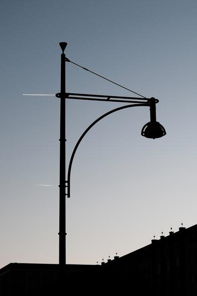 Das Foto zeigt eine Straßenlaterne vor einem wolkenlosen Abendhimmel und einem Häuserdach. Die Lampe ist nicht eingeschaltet. Die Kondensstreifen zweier Flugzeuge am Himmel erscheinen genau auf der Höhe von zwei Querstreben der Laterne. Laterne und Dach erscheinen als Silhouette. Auf dem Dach sind zwei Gruppen von je vier Schornsteinen zu sehen, die mit einem senkrechten Stab mit einer Kugel verziert sind.

The photo shows a street lamp in front of a cloudless evening sky and the roof of a house. The lamp is not switched on. The contrails of two airplanes in the sky appear exactly at the height of two cross struts of the lantern. The lantern and roof appear as a silhouette. Two groups of four chimneys can be seen on the roof, each decorated with a vertical rod with a sphere.