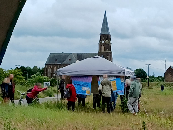 Rund um einen Zeltpavillon stehen Menschen auf einer Wiese und betrachten aufgehängte Bild-Leinwände, im Hintergrund die ehemalige Kirche von Manheim