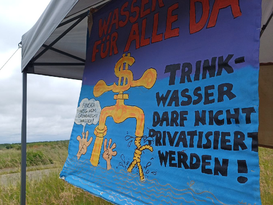 An einem grauen Zelt-Pavillon hängt eine bemalte Leinwand mit blauem Bildgrund und einem goldenen Wasserhahn mit Dollar-Zeichen, im Hintergrund Wiesen und eine Straße
