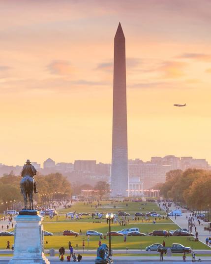 The image depicts the Washington Monument, a prominent obelisk located in Washington D.C. The monument stands tall against a backdrop of a vibrant sunset, casting a warm glow over the scene. In the foreground, there is a statue of a man on horseback, likely a historical figure. The area around the monument is bustling with people and cars, indicating a popular tourist destination. The sky is adorned with a few clouds, and an airplane can be seen flying in the distance, adding to the dynamic atmosphere of the scene. The overall ambiance is lively and picturesque, capturing a moment of everyday life at this iconic landmark.