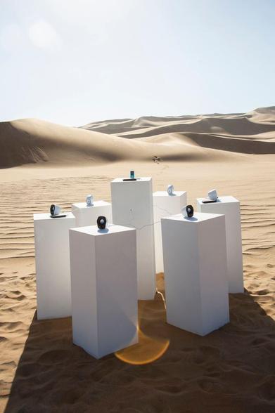 Sechs weiße Lautsprecher stehen auf rechteckigen Podesten in einer Sandwüste. Im Hintergrund sind sanfte Sanddünen sichtbar und der Himmel ist klar. Ein MP3-Player ist zwischen den Lautsprechern platziert, und einige Kabel verbinden die Lautsprecher miteinander.