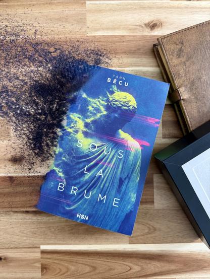 Livre posé sur une table avec un effritement de sa couverture en sable. Un bout de cadre et un vieux carnet sont posés à côté. La couverture de Sous la brume montre une statue antique dont une partie s’effrite en poussières. Des artéfacts de glyphes apparaissent aussi sur l’image.
